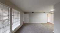 This single-floor unit offers a straightforward and functional layout spread across several connected rooms. The entryway features vertical shiplap-style wall paneling and large windows fitted with horizontal blinds, flooding the space with natural light from the moment you step inside.

The open living area is finished with neutral gray carpet and light gray walls, creating a clean, understated aesthetic. Multiple doorways connect the living space to adjacent rooms, offering a smooth flow throughout the unit. A second carpeted area, visible from the main living space, transitions to hard-surface flooring, delineating different functional zones within the home.

The galley kitchen is well-appointed with white shaker-style cabinetry lining both walls, light stone-patterned countertops, a stainless steel sink, and wood-look plank flooring. Appliances include a stainless steel refrigerator, a black range, and a dishwasher, making the kitchen both practical and visually cohesive.

The bedroom features soft carpeting, a ceiling fan with integrated lighting, a window with blinds for natural light control, and direct access to a closet and a private bathroom. The bathroom includes a stand-up shower enclosure with frosted glass doors and tiled surround, along with standard fixtures. Throughout the unit, consistent neutral tones and clean finishes contribute to a calm, move-in-ready environment.