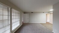This single-floor unit offers a straightforward and functional layout spread across several connected rooms. The entryway features vertical shiplap-style wall paneling and large windows fitted with horizontal blinds, flooding the space with natural light from the moment you step inside.

The open living area is finished with neutral gray carpet and light gray walls, creating a clean, understated aesthetic. Multiple doorways connect the living space to adjacent rooms, offering a smooth flow throughout the unit. A second carpeted area, visible from the main living space, transitions to hard-surface flooring, delineating different functional zones within the home.

The galley kitchen is well-appointed with white shaker-style cabinetry lining both walls, light stone-patterned countertops, a stainless steel sink, and wood-look plank flooring. Appliances include a stainless steel refrigerator, a black range, and a dishwasher, making the kitchen both practical and visually cohesive.

The bedroom features soft carpeting, a ceiling fan with integrated lighting, a window with blinds for natural light control, and direct access to a closet and a private bathroom. The bathroom includes a stand-up shower enclosure with frosted glass doors and tiled surround, along with standard fixtures. Throughout the unit, consistent neutral tones and clean finishes contribute to a calm, move-in-ready environment.