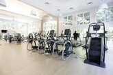 Located at 13147–13151 Morris Rd in Milton, Georgia, this fitness center offers a bright, expansive workout environment designed to support a comprehensive exercise experience. The single-floor facility is characterized by soaring ceilings, large multi-pane windows, and an abundance of natural light that floods the open interior throughout the day. The views from nearly every vantage point overlook lush, mature greenery, creating a serene and motivating atmosphere.

The main workout floor is finished with light hardwood-style flooring and organized into distinct zones. The cardio section is well-appointed with multiple Matrix treadmills, elliptical trainers, and stationary bikes, all positioned to take full advantage of the floor-to-ceiling windows and wooded outdoor views. A dedicated strength training area features a broad selection of professional-grade cable machines, weight benches, and selectorized equipment, complemented by wall-mounted mirrors that enhance the sense of openness.

A separate flex area provides space for functional training and stretching, outfitted with an adjustable bench and stability ball. Pendant lighting and ceiling fans contribute to a clean, polished aesthetic throughout. The facility's architecture blends traditional detailing—including white-trimmed columns and paneled ceilings—with a modern, airy design that makes this fitness center both functional and visually inviting.