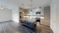 This single-floor residence showcases a thoughtfully designed contemporary interior with a cohesive modern aesthetic throughout. The kitchen is a standout feature, equipped with stainless steel appliances including a French door refrigerator, electric range, and over-the-range microwave. Two-toned cabinetry pairs glossy white upper cabinets with warm wood-grain lower cabinets, complemented by white quartz countertops, a subway tile backsplash accented by a decorative mosaic border, and wood-look flooring. Pendant lighting illuminates the space effectively.

The living area is bright and welcoming, anchored by large sliding glass doors that open to a private balcony with urban views. A ceiling fan, dark accent wall, and wood-look flooring define the space's modern character. The bedrooms feature plush carpeting, ceiling fans, and windows that invite natural light.

Two bathrooms serve the unit: a primary bath with a glass-enclosed walk-in shower finished in white tile with a mosaic accent band, and a secondary bath with light tile flooring and a floating dark-toned vanity. Neutral wall tones and clean lines maintain a consistent, contemporary feel throughout the entire residence.