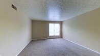 Located at 4388 Walford Street in Columbus, Ohio, this single-floor unit offers a straightforward and functional layout designed for comfortable everyday living. The main living area is an open space with speckled carpet flooring and neutral beige walls that create a clean, understated aesthetic. A textured popcorn ceiling runs throughout, and baseboard heating units are positioned along the walls, providing warmth throughout the colder months. A partial wall divider subtly separates the living space from the adjacent kitchen area, which features white cabinetry and a white refrigerator, offering a practical cooking and storage setup. A doorway from the living area leads further into the unit. A second carpeted room, likely a bedroom, offers a quieter retreat with its own baseboard heater and a sliding window that allows natural light to fill the space. The neutral color palette and simple finishes throughout give the unit a blank-canvas quality, well-suited for personalization. Overall, this unit presents a no-frills, functional living environment in a convenient Columbus location.