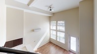 This two-story space offers a thoughtfully designed layout with contemporary finishes throughout. The main living area is bright and airy, benefiting from soaring ceilings, warm hardwood-style flooring, and a bank of large windows that flood the space with natural light. A ceiling fan adds both comfort and style to the upper portion of the room, while a wall-mounted air conditioning unit ensures year-round climate control. A sleek, modern staircase with a black steel frame and horizontal cable railing connects the two levels, serving as a striking architectural focal point. The kitchen is efficiently designed, featuring dark espresso cabinetry, white countertops, a classic subway tile backsplash, and a full suite of stainless steel appliances including a refrigerator, electric range, over-the-range microwave, and dishwasher. The bedroom, visible from the upper vantage point, is carpeted for comfort and includes a spacious closet with double doors. The bathroom is clean and functional, offering a full bathtub and shower combination, wood-style flooring, and accessibility grab bars. Neutral wall tones and consistent flooring throughout create a cohesive and inviting atmosphere across both levels.