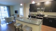 This single-floor residence features an open-concept layout that seamlessly connects the kitchen and living area. The kitchen is a standout, showcasing a large granite-topped island with an undermount sink, bar seating for two, and dual pendant lighting overhead. Dark espresso cabinetry contrasts elegantly against light-toned walls and a classic subway tile backsplash. Stainless steel appliances, including a range, microwave, and dishwasher, complete the modern kitchen setup. Warm hardwood flooring extends throughout the space, adding a cohesive and inviting feel. The adjacent living area is visible from the kitchen, featuring a wall-mounted flat-screen TV and a low-profile storage unit, creating a comfortable and functional gathering space. A window allows natural light to filter into the area, brightening the open floor plan. The overall design balances contemporary finishes with a welcoming ambiance, making the space well-suited for both everyday living and entertaining.