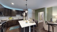 This two-bedroom, two-bathroom single-floor residence delivers a thoughtfully designed open-concept living space. The kitchen is a standout feature, showcasing dark espresso cabinetry paired with a clean white tile backsplash and stainless steel appliances. A large kitchen island with a bright white countertop serves as both a prep space and casual dining area, complemented by stylish pendant lighting that hangs overhead, adding warmth and visual interest. Rich dark hardwood-style flooring flows throughout, creating a cohesive and sophisticated aesthetic. An accent wall painted in a soft sage green adds a touch of color and depth to the living and dining area, while framed artwork enhances the overall ambiance. The open layout allows for a natural transition between the kitchen, dining, and living spaces, contributing to a spacious and functional feel. The single-floor design ensures easy accessibility throughout. Overall, the residence combines contemporary design elements — including bold cabinetry contrasts, modern lighting fixtures, and warm flooring tones — to create a stylish and comfortable living environment suited for everyday life.