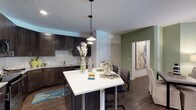 This two-bedroom, two-bathroom single-floor residence delivers a thoughtfully designed open-concept living space. The kitchen is a standout feature, showcasing dark espresso cabinetry paired with a clean white tile backsplash and stainless steel appliances. A large kitchen island with a bright white countertop serves as both a prep space and casual dining area, complemented by stylish pendant lighting that hangs overhead, adding warmth and visual interest. Rich dark hardwood-style flooring flows throughout, creating a cohesive and sophisticated aesthetic. An accent wall painted in a soft sage green adds a touch of color and depth to the living and dining area, while framed artwork enhances the overall ambiance. The open layout allows for a natural transition between the kitchen, dining, and living spaces, contributing to a spacious and functional feel. The single-floor design ensures easy accessibility throughout. Overall, the residence combines contemporary design elements — including bold cabinetry contrasts, modern lighting fixtures, and warm flooring tones — to create a stylish and comfortable living environment suited for everyday life.