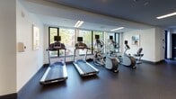 Located at 1478 Tremont St in Boston's Mission Hill neighborhood, this fitness center offers a comprehensive, modern workout facility designed to support a full range of exercise routines. The space is thoughtfully organized into distinct training zones, all united by a clean, contemporary aesthetic with white walls, dark rubberized flooring, and recessed linear lighting overhead.

The cardio area is lined with large windows that frame views of surrounding greenery, flooding the space with natural light while providing an energizing backdrop for treadmills and elliptical machines, each equipped with individual display screens. A dedicated strength and functional training zone features full-length mirrors that amplify the sense of space, alongside a multi-station cable machine, a flat workout bench, and a well-stocked rack of dumbbells ranging across multiple weight increments. A functional training station rounds out the equipment selection with stability balls in assorted sizes, resistance bands, exercise mats, and medicine balls organized on a dedicated storage rack. A posted "Tremont Fitness Rules" placard near the entrance reinforces the facility's professional, community-oriented environment. The single-floor layout ensures seamless flow between all training areas, making this fitness center a well-equipped and accessible amenity.