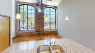 This one-bedroom, one-bathroom unit in Charlotte's Plaza Midwood area blends industrial character with modern finishes. The open living and kitchen area showcases soaring ceilings, original exposed brick walls, large arched factory-style windows, and warm hardwood floors that flood the space with natural light. The kitchen features granite countertops, rich dark wood cabinetry, stainless steel appliances, and stylish pendant lighting. The bedroom offers a sliding wood door, built-in closet with wire shelving, and the same hardwood flooring. The clean bathroom includes a tiled floor, tub/shower combo, and granite-topped vanity.