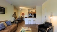 This single-story residence showcases an open-concept floor plan that integrates the living, dining, and kitchen areas into one cohesive, flowing space. The warm hardwood-style flooring runs continuously throughout, anchoring the layout and lending a sense of warmth and continuity.

The living area is generously proportioned, accommodating a comfortable seating arrangement with ample room to move. It transitions naturally into the dining area, which features a dedicated dining set and is accented by a decorative wall niche—an architectural detail that adds character to the space.

The kitchen is positioned at the rear of the open layout and is defined by a breakfast bar with bar stool seating, offering both functionality and a casual dining option. Medium-toned wood cabinetry lines the kitchen walls, complemented by a light granite-style countertop and black appliances, including a refrigerator, stove, and microwave. A pass-through design keeps the kitchen visually connected to the rest of the living space.

Crown molding trims the upper walls, adding a refined touch, while track lighting illuminates the main living area. The overall ambiance is warm, inviting, and well-suited for both everyday living and casual entertaining.