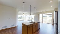 This single-floor space is designed with an open-concept layout that seamlessly connects the kitchen and living areas, creating a spacious and airy atmosphere. The kitchen serves as the heart of the space, anchored by a generously sized center island topped with a light-toned countertop and equipped with a double stainless steel sink and a built-in dishwasher. Warm wood cabinetry with sleek hardware adds a natural, inviting tone to the kitchen. Three pendant lights hang from the ceiling on staggered black cords, providing focused task lighting over the island while contributing a modern design element.

The open living area beyond the kitchen benefits from an abundance of natural light, courtesy of large floor-to-ceiling windows that frame unobstructed views of the surrounding green landscape. A tray ceiling detail adds architectural interest to the living space. Track lighting along the ceiling offers additional flexibility. Rich hardwood flooring flows throughout, grounding the space with warmth and continuity.

A private balcony accessible from the main living area extends the space outdoors, offering panoramic views of open fields and a quiet road under wide, open skies—ideal for enjoying the natural surroundings.