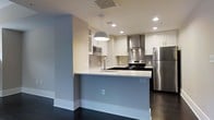 This two-floor space showcases a clean, contemporary design with a thoughtful layout and modern finishes throughout. The kitchen is a standout feature, offering a peninsula-style configuration that creates a natural division between the cooking and living areas. White shaker-style cabinetry lines the walls, complemented by a metallic tile backsplash and a stainless steel range hood that adds an industrial-chic accent. Stainless steel appliances — including a refrigerator, electric range, and built-in microwave — provide a cohesive, modern aesthetic. The white countertops offer ample prep space, and a stylish pendant light hangs above the peninsula, adding a warm focal point to the room. Recessed lighting throughout ensures the space is well-lit, while dark hardwood flooring grounds the light-toned walls and cabinetry with a rich contrast. The neutral color palette of soft grays and whites creates a bright, airy ambiance that feels both inviting and functional. The second floor adds additional living space, expanding the overall versatility of this well-designed property. The overall atmosphere is modern, clean, and move-in ready.