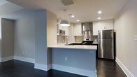 This two-floor space showcases a clean, contemporary design with a thoughtful layout and modern finishes throughout. The kitchen is a standout feature, offering a peninsula-style configuration that creates a natural division between the cooking and living areas. White shaker-style cabinetry lines the walls, complemented by a metallic tile backsplash and a stainless steel range hood that adds an industrial-chic accent. Stainless steel appliances — including a refrigerator, electric range, and built-in microwave — provide a cohesive, modern aesthetic. The white countertops offer ample prep space, and a stylish pendant light hangs above the peninsula, adding a warm focal point to the room. Recessed lighting throughout ensures the space is well-lit, while dark hardwood flooring grounds the light-toned walls and cabinetry with a rich contrast. The neutral color palette of soft grays and whites creates a bright, airy ambiance that feels both inviting and functional. The second floor adds additional living space, expanding the overall versatility of this well-designed property. The overall atmosphere is modern, clean, and move-in ready.