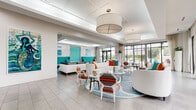 Located at 4460 Lower Park Rd in Orlando's Baldwin Park neighborhood, Azul is a single-story clubhouse facility designed with a clean, contemporary aesthetic. The space makes an immediate impression at the reception area, where a sleek white desk with frosted glass panels and the "AZUL" nameplate in metallic lettering sets an inviting, polished tone. Large-format light gray tile flooring flows throughout, lending a cohesive and modern feel across the entire floor plan. The main lounge is expansive and open, filled with abundant natural light streaming through floor-to-ceiling black-framed glass walls and doors. The interior is furnished with a curated mix of white sofas, gray sectionals, and bold teal tufted banquettes accented by vibrant orange and multicolored throw pillows, creating an energetic yet sophisticated atmosphere. Abstract ocean-inspired artwork adorns the marble-look accent walls, reinforcing the coastal color palette. Coffered ceilings with recessed lighting and large drum pendant fixtures add architectural interest overhead. The space flows seamlessly toward the pool area through expansive sliding glass doors and French doors, offering unobstructed views of the outdoor pool deck, lounge chairs, and palm trees beyond—bringing the Florida sunshine indoors. Multiple distinct seating zones are arranged throughout, accommodating both social gatherings and relaxed lounging.