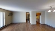 This single-floor residence offers a clean, functional layout designed for comfortable everyday living. The main living area is open and airy, featuring smooth white ceilings and warm wood-style laminate flooring that extends throughout the space, lending a cohesive and inviting feel. Neutral-toned walls provide a versatile backdrop, making the space easy to personalize.

A hallway leads from the living area to additional rooms, with natural light visible at the end, suggesting well-placed windows that help brighten the interior. The kitchen is tucked to the side in a semi-open configuration, offering warm medium-toned wood cabinetry and a full suite of white appliances, including a refrigerator and stovetop. Under-cabinet lighting adds a practical touch and enhances the kitchen's warm ambiance.

The overall layout is straightforward and efficient, making good use of the available floor plan. The consistent flooring, neutral palette, and clean lines throughout create a tidy, well-maintained atmosphere. This space is well-suited for those seeking a simple, move-in-ready residence with a practical design and a welcoming, understated aesthetic.