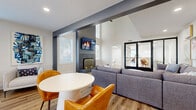 Located at 303 Sutton Cir in Daytona Beach, Florida, this well-appointed single-floor community space offers a thoughtfully designed interior that blends modern aesthetics with comfortable functionality. The expansive open-plan living area is the heart of the space, anchored by a dramatic floor-to-ceiling shiplap fireplace surround in charcoal gray, fitted with a gold-accented hearth and a wall-mounted television above. Soaring ceilings with exposed dark beams add architectural character, while wide-plank wood-look flooring runs throughout, lending warmth and visual continuity. Multiple seating arrangements—including a generous sectional sofa, lounge chairs, and round café-style tables with vibrant amber leather chairs—create versatile zones for relaxing or socializing. Recessed lighting and a sleek ceiling fan provide comfort, while abundant natural light pours in through numerous windows dressed with clean white blinds and glass-paned double doors leading to adjacent areas. A built-in white credenza offers additional functionality near a dedicated beverage station. A glass-walled private office or meeting room adds a professional touch. The bathroom features light wood-look flooring, a pedestal sink, and a minimalist round mirror. Curated coastal artwork throughout enhances the bright, welcoming ambiance of this Florida retreat.