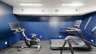 Located at 7502 Hunt Club Rd in Columbia, South Carolina, this dedicated fitness room offers a focused and functional workout environment within a compact, single-floor layout. The space is immediately defined by its bold navy blue walls, which extend from a dark base up to a clean white ceiling, lending the room a high-energy, gym-like atmosphere. Durable speckled gray flooring provides a practical, easy-to-maintain surface well-suited for exercise equipment and active use.

The room is equipped with commercial-grade Matrix fitness machines, including a stationary cycle, a treadmill, and a robust multi-functional cable machine — providing users with a comprehensive range of cardiovascular and strength-training options. Overhead fluorescent lighting ensures the space is consistently and evenly lit, supporting safe use of all equipment at any time of day.

Two doors serve the room: a white paneled exterior door with frosted sidelights providing access from outside, and a classic French-style interior door with divided glass panes connecting to an adjacent area. An exit sign and security camera are prominently mounted near the entry, underscoring the space's attention to safety. Signage on the walls reinforces community guidelines, including restrictions on alcoholic beverages and loud music, indicating this is a shared amenity space.