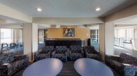 This single-floor communal space is thoughtfully designed to accommodate a variety of activities, from casual socializing to focused group meetings. The open layout is anchored by patterned carpet in deep blue and teal tones, which runs throughout and ties the various zones together cohesively.

The main lounge area features multiple patterned upholstered armchairs arranged around round tables, creating an inviting setting for conversation or group work. A built-in wet bar with dark cabinetry and a sink lines one wall, accented by a vibrant piece of abstract wall art that adds a splash of color to the neutral beige surroundings.

Along the perimeter, a sunlit corridor zone is furnished with high-top tables and modern bar-height chairs in bright teal, bathed in natural light from large windows that overlook the exterior. This area offers a more informal, energetic atmosphere suited for quick meetings or individual work.

A dedicated meeting alcove includes a rectangular table with chairs and a large window fitted with vertical blinds, providing controllable natural light and a quieter setting for focused collaboration.

The textured ceiling, recessed lighting, and structured column details throughout lend the space a practical yet polished character, making it well-suited for use as a student lounge, common room, or shared community gathering area.