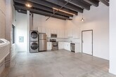 This single-floor space showcases a compelling industrial aesthetic defined by soaring ceilings with bold, dark-stained exposed wood beams and polished concrete floors throughout the main living area. The open layout creates a generous sense of space and volume, with pendant lighting and a linear track fixture adding warm, focused illumination beneath the dramatic ceiling structure.

The kitchen is well-appointed with white shaker-style cabinetry extending to the upper walls, taking full advantage of the elevated ceiling height for maximum storage. A complete suite of stainless steel appliances — including a refrigerator, electric range, built-in microwave, and dishwasher — provides all modern conveniences. Light-toned countertops complement the clean, crisp cabinetry. A stacked stainless steel washer and dryer unit is conveniently located adjacent to the kitchen area.

The living space features exposed concrete block walls that reinforce the raw, urban character of the unit, along with a window that frames views of the surrounding neighborhood and allows natural light to fill the room.

The bathroom is finished in a clean, minimalist style with white walls, a vanity featuring a light stone-style countertop, white cabinetry, and a full tub/shower combination. Stone-look tile flooring adds a subtle texture to the space. The overall ambiance is modern and utilitarian, ideal for those who appreciate an urban loft aesthetic.