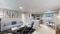 This single-floor residence offers a thoughtfully designed open-concept layout that seamlessly connects the living area and kitchen, creating a cohesive and functional living space. Light wood-style flooring extends throughout the main living areas, complementing the neutral gray-toned walls and contributing to a bright, modern aesthetic.

The living area is generously proportioned, featuring a large window that invites natural light and provides views of the surrounding exterior. A front entry door opens directly into this space, which comfortably accommodates seating arrangements alongside a decorative credenza and accent furnishings.

The kitchen is well-equipped with gray cabinetry, white countertops, a subway tile backsplash, and a full suite of stainless steel appliances, including a refrigerator, electric range, and over-the-range microwave. A peninsula with an integrated sink offers additional prep space and separates the kitchen from the living area.

The bedroom features plush carpet flooring, a window with natural light, and ample wall space. Two bathrooms serve the unit, each finished with gray cabinetry, white countertops, wood-look flooring, and a tiled tub-shower combination.