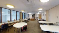 This single-floor multipurpose space is designed to accommodate a variety of activities, from meetings and study sessions to community gatherings. The room is furnished with multiple round and rectangular white tables paired with folding chairs, allowing for flexible seating arrangements to suit different group sizes and configurations. Along one wall, a continuous bank of large windows allows generous natural light to filter into the space, creating a bright and welcoming atmosphere. The drop ceiling is fitted with a combination of flush-mounted circular light fixtures and recessed fluorescent panels, ensuring consistent illumination throughout. A wall-mounted flat-screen television serves as a focal point at one end of the room, making it well-suited for presentations or group viewings. A wooden bookshelf unit stocked with books and media adds a resourceful, library-like element to the space. Neutral carpeted flooring and light-colored walls contribute to a calm, functional ambiance. A standard interior door provides access to an adjacent area, and framed artwork on the walls adds a modest decorative touch. Overall, the space is practical, organized, and adaptable for a wide range of uses.
