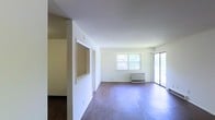 This single-floor space presents an open, functional layout defined by clean white walls and warm hardwood flooring throughout. The main living area is generously sized, allowing for flexible furniture arrangements to suit various needs. Natural light fills the room through a pair of windows along the far wall, complemented by a sliding glass door that provides additional brightness and direct outdoor access, enhancing the overall sense of openness.

A wall-mounted HVAC unit is positioned beneath the windows, providing climate control directly within the living space. A distinctive architectural detail is the pass-through window cutout built into the interior wall, connecting adjacent areas and contributing to an open, communicative flow between spaces. This feature, combined with the open sightlines, gives the interior a connected and airy feel.

The adjoining area, visible through the pass-through and a doorway, features a slightly different flooring tone, suggesting a defined transition between functional zones. The neutral color palette throughout creates a blank canvas, well-suited for a variety of interior styles. Overall, the space offers a straightforward, practical layout with key features that support comfortable, everyday living.
