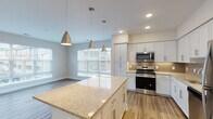This single-floor residence is designed with a clean, contemporary aesthetic that balances functionality and style throughout. The open-concept kitchen serves as the heart of the home, showcasing white shaker cabinetry extending to the ceiling, granite countertops, and a large central island ideal for casual dining or meal preparation. A trio of brushed gold pendant lights hangs above the island, complementing the stainless steel appliances, which include a gas range, built-in microwave, dishwasher, and refrigerator. A subtle tile backsplash adds texture behind the range and sink area, while natural light pours in through large windows overlooking the adjacent dining and living space.

The open living area continues the warm hardwood-style flooring, providing a generous, unobstructed layout. Bedrooms are carpeted for comfort and include ample closet space with built-in rod systems. Two bathrooms serve the residence: a full bath featuring white subway tile surrounding a soaking tub, a granite vanity, and tile flooring, and a second bathroom with a matching granite vanity and tile floor. The overall palette of soft neutrals, white trim, and brushed hardware creates a cohesive and inviting atmosphere throughout.