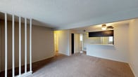 This single-floor space presents an open-concept layout that seamlessly connects the living area to the kitchen. The living space is finished with neutral-toned wall-to-wall carpeting, creating a warm and cohesive atmosphere throughout. A textured popcorn ceiling runs the length of the main area, adding a classic residential character.

The kitchen is positioned at the rear of the open layout and features dark upper cabinetry paired with a light-colored countertop that extends into a pass-through or breakfast bar, offering a functional divide between the cooking and living spaces. A ceiling fan with a built-in light fixture illuminates the kitchen area, providing both ventilation and ambient lighting.

From the main living area, multiple doorways lead to additional rooms, suggesting a practical and accessible floor plan. One hallway reveals what appears to be a closet with sliding doors, while another opening leads further into the unit, hinting at bedroom or bathroom access. The neutral wall tones throughout complement the carpeted floors, giving the space a clean, move-in-ready appearance.