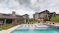 This residential community showcases a thoughtfully designed outdoor living space centered around a stunning resort-style swimming pool. The pool area features in-water lounge chairs and sun shelves, ideal for relaxation, surrounded by a spacious concrete deck lined with sleek chaise lounges and potted ornamental grasses and tropical plantings that add a lush, resort-like atmosphere.

The architecture of the surrounding buildings blends a mix of classic brick, stone, and modern board-and-batten siding in neutral tones of gray and tan, complemented by pitched rooflines and large windows that allow abundant natural light into the interiors. Multi-story residential buildings frame the pool courtyard, offering elevated balconies and views of the amenity area below.

Adjacent to the pool, a dedicated clubhouse or amenity building with expansive glass windows provides an indoor-outdoor connection to the pool deck, while a separate outdoor dining and gathering area equipped with shaded seating and an umbrella can be seen in the background. Decorative stonework retaining walls and carefully curated landscaping with native grasses and palm trees tie the outdoor spaces together seamlessly, creating a cohesive and welcoming community environment.