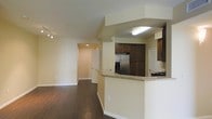 This single-floor residence offers a thoughtfully designed open-concept layout that seamlessly connects the kitchen and living areas. The kitchen is a standout feature, showcasing rich dark wood cabinetry that extends to the upper walls, offering generous storage space. A granite-topped breakfast bar serves as a natural divider between the kitchen and the living area, providing both a casual dining option and an elegant visual anchor. Stainless steel appliances, including a French door refrigerator with an ice and water dispenser, add a modern, functional touch to the kitchen. A gas cooktop is also visible, catering to culinary enthusiasts. The living area flows openly from the kitchen, bathed in warm, neutral tones on the walls that complement the rich, dark hardwood flooring running throughout the space. Track lighting in the living area provides flexible illumination, while recessed lighting in the hallway adds a polished finishing touch. The hallway leads further into the residence, hinting at additional private spaces beyond. Bright white trim and baseboards provide clean contrast throughout, contributing to a cohesive and well-maintained aesthetic. Overall, the space balances practicality with modern style.