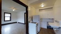 This single-floor space showcases a blend of classic architectural character and functional layout. The kitchen is equipped with white cabinetry, a gas range, and a full-sized refrigerator, with upper and lower cabinets providing ample storage. A flush-mount ceiling light illuminates the kitchen area, which features a darker tile flooring that contrasts with the lighter tones of the cabinetry and walls. Crown molding accents the upper edges of the walls, adding a traditional architectural detail throughout.

A wide, dark-trimmed arched doorway connects the kitchen to an adjoining living area, creating an open and accessible flow between the two spaces. The living room features speckled terrazzo-style flooring and its own ceiling fan, contributing to a practical and airy feel. Crown molding continues into this room, maintaining design consistency. A hallway visible through the living area suggests additional rooms beyond. The neutral wall tones throughout keep the interior feeling bright and cohesive, while the classic trim work lends the space a sense of enduring architectural character.