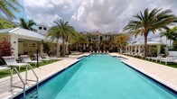 This stunning resort-style amenity space showcases an impressively sized rectangular swimming pool as its centerpiece, finished with vibrant blue tile that reflects the Florida sky above. The pool is framed by a broad, light-toned paver deck generously lined with cushioned lounge chairs, offering ample space for sunbathing and relaxation. Towering royal palm trees are strategically placed throughout the grounds, providing both shade and a distinctly tropical ambiance. Lush tropical plantings, including bursts of red flowering shrubs, add color and visual depth to the landscape. The surrounding architecture is elegant and refined, featuring a multi-story building with balconies, classic columns, and a sleek metal roof that blends Florida vernacular style with upscale residential design. Covered lounge areas and pergola-style structures line the perimeter, offering shaded retreats. The overall atmosphere is bright, open, and inviting, evoking the feel of a high-end resort. The combination of manicured grounds, expansive hardscape, and lush greenery creates a cohesive and polished outdoor environment designed for leisure and community enjoyment.
