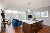 This single-floor residence offers a thoughtfully designed open-concept layout that seamlessly connects the kitchen, dining, and living areas. The kitchen is a standout feature, showcasing rich walnut-toned cabinetry, stainless steel appliances, a tile backsplash, and a generously sized island with granite countertops and bar seating — ideal for both everyday meals and entertaining. Track and pendant lighting add functional ambiance overhead, while light hardwood flooring flows consistently throughout the space.

The living area is bathed in natural light through expansive floor-to-ceiling sliding glass doors that open to an outdoor balcony, offering views of surrounding trees and the neighborhood beyond. A long, art-lined hallway connects the main living area to the private quarters, adding a gallery-like quality to the interior.

The bedroom is a calm retreat, anchored by a neutral accent wall and ample space for furnishings. Adjacent to the bedroom is a well-organized walk-in closet with built-in shelving and hanging storage, accented by a decorative gold mirror and side table. The bathroom features a clean, minimalist design with marble-toned countertops, a shower with a classic white curtain, and tasteful wall art, completing this refined and functional home.