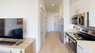 Located on East 6th Street in Austin's vibrant 78702 neighborhood, this modern single-floor unit offers a thoughtfully designed 2-bedroom, 1-bathroom layout that maximizes every square foot. Light-toned hardwood-style flooring runs throughout, tying together a clean, contemporary aesthetic across all rooms.

The galley-style kitchen is well-appointed with stainless steel appliances—including a refrigerator, range, and over-the-range microwave—complemented by white cabinetry extending to the ceiling, light quartz countertops, and a grey tile backsplash. A hallway with open shelving connects the kitchen to the main living area, offering practical built-in storage.

Both bedrooms feature crisp white walls, ceiling fans, and windows with blinds and bold patterned curtains that allow for natural light control. The primary bedroom accommodates a queen bed and includes a full-length mirror, while the second bedroom mirrors the same modern, minimalist design with multiple windows providing ample brightness.

The bathroom is clean and functional, finished with wood-look tile flooring, a tub-shower combination with a tiled surround featuring a decorative accent stripe, and upper storage cabinetry. Curated artwork is displayed throughout the unit, adding warmth and personality to the otherwise neutral palette.