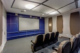 Located at 8610 N Kansas Ave in Kansas City, Missouri, this purpose-built screening room delivers a true cinematic experience within a single-floor layout. The space is anchored by a large projection screen mounted along a navy blue accent wall, framed by built-in shelving units that line the front of the room, adding both functionality and a polished aesthetic.

Rows of upholstered theater-style seats with tan fabric and black frames are arranged in a tiered configuration, ensuring clear sightlines to the screen from every position. Each seat features integrated armrests with cup holders, designed for comfort during extended viewing sessions.

Acoustic performance is a defining characteristic of this room. Numerous large, oval-shaped sound absorption panels are mounted across the side and rear walls in a warm dark brown tone, effectively controlling sound reflections and optimizing audio clarity. The ceiling continues this acoustic design with a coffered grid of panels, complementing the controlled sound environment.

The flooring throughout is a durable, multi-tone commercial carpet in blue-grey tones, and recessed and panel lighting provide even, adjustable illumination. A double-door entry with electronic access control is positioned at the rear of the room. Overall, the space is thoughtfully designed to function as a professional-grade screening and presentation venue.