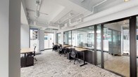 Located on Pacific Avenue in downtown Long Beach, California, this single-floor coworking space offers a contemporary and highly functional work environment designed to support a range of professional needs. The layout combines open-plan workstations with glass-walled private offices, providing flexibility for both collaborative and focused individual work.

The open workspace features rows of natural wood-topped, height-adjustable desks paired with ergonomic black task chairs, all set atop a textured, patterned carpet that adds visual warmth to the otherwise clean, industrial aesthetic. Exposed white ductwork and conduit run along the ceiling, complemented by track lighting that ensures consistent illumination throughout the space.

Full-height glass partitions with dark metal frames divide the open area from enclosed private offices, maintaining visual connectivity while offering acoustic separation. These private offices are furnished with individual desks and chairs, suitable for focused work or small meetings.

Floor-to-ceiling street-facing windows flood the space with natural light and offer direct views of the surrounding downtown Long Beach streetscape, including tree-lined sidewalks and nearby urban architecture. The transparent façade creates an open, energizing atmosphere well-suited to productive workdays in the heart of the city.