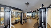 This fitness center is designed to accommodate a wide range of exercise needs within a single, efficiently organized space. The layout is defined by a striking curved glass-and-dark-frame enclosure near the entrance, which separates a dedicated workout zone while maintaining visual openness throughout the facility. The flooring is finished in a durable, cushioned rubberized material in a neutral speckled tone, providing both comfort and safety during exercise.

The main floor area is outfitted with a selection of strength training equipment, including a weight bench and adjustable resistance machines, as well as medicine balls for functional training. Large wall-mounted mirrors extend across one side of the room, visually expanding the space and allowing users to monitor their form during workouts. Recessed lighting and overhead ventilation ensure a comfortable environment throughout.

Beyond the primary exercise area, additional equipment is visible deeper in the space, suggesting room for cardio machines or supplementary training stations. Sight lines through the glass walls reveal an adjacent area, possibly a lounge or amenity room, contributing to a connected and transparent overall atmosphere within the facility.