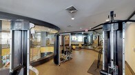 This fitness center is designed to accommodate a wide range of exercise needs within a single, efficiently organized space. The layout is defined by a striking curved glass-and-dark-frame enclosure near the entrance, which separates a dedicated workout zone while maintaining visual openness throughout the facility. The flooring is finished in a durable, cushioned rubberized material in a neutral speckled tone, providing both comfort and safety during exercise.

The main floor area is outfitted with a selection of strength training equipment, including a weight bench and adjustable resistance machines, as well as medicine balls for functional training. Large wall-mounted mirrors extend across one side of the room, visually expanding the space and allowing users to monitor their form during workouts. Recessed lighting and overhead ventilation ensure a comfortable environment throughout.

Beyond the primary exercise area, additional equipment is visible deeper in the space, suggesting room for cardio machines or supplementary training stations. Sight lines through the glass walls reveal an adjacent area, possibly a lounge or amenity room, contributing to a connected and transparent overall atmosphere within the facility.