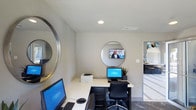 This Dallas property features a well-appointed business center designed to support productivity in a polished, contemporary setting. The single-story space showcases a clean, modern aesthetic with white walls, recessed lighting, and a neutral color palette that creates a bright and focused atmosphere.

The workstation area is anchored by an L-shaped white desk offering ample surface space for multiple users simultaneously. Two computer workstations with monitors and keyboards are set up for convenient use, complemented by a printer, making the space fully equipped for business tasks. A sleek black ergonomic office chair provides comfortable seating at the primary station.

Decorative round mirrors with silver metallic frames are mounted on the walls, adding a stylish, contemporary touch while enhancing the sense of openness in the room. A wall-mounted flat-screen television is positioned above one of the mirrors, adding an entertainment or informational element to the space.

An open doorway leads to an adjacent area with lighter cabinetry and countertops visible in the background, suggesting easy access to additional amenities. The overall design balances functionality with modern style, making it a practical and inviting workspace.
