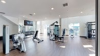This contemporary fitness center spans two floors, offering a comprehensive and well-appointed workout environment designed to support a wide range of exercise routines. The space is characterized by a bright, clean aesthetic, featuring white walls, recessed lighting, and durable wood-style flooring throughout.

The main fitness floor is generously proportioned and houses an extensive selection of cardio equipment, including multiple treadmills, elliptical trainers, stair climbers, and stationary bikes — all equipped with individual screens for an engaging workout experience. Large windows and glass railings line the perimeter, allowing abundant natural light to fill the space and offering views of the outdoor surroundings, including what appears to be a pool area.

A dedicated strength training zone features a multi-station cable machine, adjustable benches, and a full rack of free weights, providing everything needed for a complete resistance training session. A wall-mounted flat-screen television adds to the amenity-rich environment.

A separate functional training studio offers a more focused space for dynamic workouts, equipped with a comprehensive rig system holding resistance bands, suspension trainers, medicine balls, kettlebells, and plyo boxes. This room features a sliding glass door that opens to natural light and exterior views, creating an energizing atmosphere for high-intensity training.