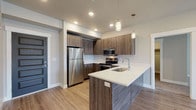 This contemporary single-floor residence showcases a thoughtfully designed open-concept layout that seamlessly connects the kitchen and living areas. The kitchen is a standout feature, outfitted with wood-grain cabinetry, a full suite of stainless steel appliances including a refrigerator, range, and over-the-range microwave, and a white quartz peninsula with an undermount sink. Pendant lighting above the peninsula adds a stylish touch, while recessed lighting throughout ensures a bright, welcoming atmosphere. Light wood-tone luxury vinyl plank flooring runs throughout the main living areas, contributing to a clean, modern aesthetic. The living area is generously proportioned and benefits from natural light streaming through a set of French doors. The bedrooms are carpeted for comfort, with neutral-toned walls and windows fitted with blinds. A walk-in closet with wrap-around shelving and hanging rods provides ample storage. The bathrooms are finished consistently with the rest of the unit, featuring wood-grain vanities with white quartz countertops, undermount sinks, large mirrors, and a tub/shower combination. Light vinyl plank flooring in the bathrooms ties the design together cohesively throughout the space.