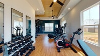 This single-floor fitness center offers a comprehensive workout environment designed to accommodate a wide range of exercise routines. The space is defined by its striking architectural ceiling detail — a tray ceiling finished with warm wood planking and fitted with ceiling fans — which adds both character and comfort to the room. Recessed lighting throughout ensures the space remains bright and functional at all times.

The flooring consists of smooth hardwood-style planks that run the length of the facility, providing a clean and durable surface. Bold navy-blue accent walls contrast with crisp white trim and walls, creating an energizing yet polished atmosphere. Multiple windows along the perimeter flood the interior with natural light and offer views of the surrounding exterior.

The fitness center is thoughtfully divided into functional zones. The cardio area houses multiple SportsArt treadmills, a StairMaster elliptical, and a Spirit exercise bike, all positioned near windows to take advantage of natural light. On the opposite side, a full dumbbell rack lines the wall alongside a large floor-length mirror, while a multi-station cable machine anchors the strength training zone. An adjustable workout bench near the entrance provides additional versatility. The space is accessed through a glass-panel door, reinforcing its open and welcoming character.