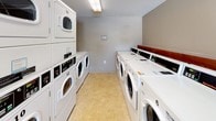 This shared laundry facility is designed to accommodate a high volume of use with efficiency and comfort in mind. The room is divided into two functional zones: a washing and drying area and a waiting and folding area. The laundry section houses a generous lineup of front-loading commercial washing machines along one wall, complemented by stacked dryer units on the opposite side, each clearly numbered for easy identification. The machines are arranged to maximize floor space while allowing comfortable movement between them. The waiting and folding area features a large L-shaped laminate folding counter that runs along the wall, providing ample surface space for sorting and folding laundry. A row of modern black chairs is positioned near the windows, offering a comfortable spot to wait. Natural light filters in through several windows, brightening the neutral-toned walls and light vinyl flooring. A self-service payment kiosk is conveniently located near the entrance, streamlining the payment process. Overhead fluorescent lighting ensures the room is well-lit throughout. Posted signage outlines the rules for machine use, and a smoke detector and fire alarm are visible, reflecting attention to safety. The textured ceiling and clean, utilitarian finishes give the space a practical and well-maintained character.