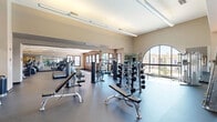 Located at 2000 Villa Dr in Pittsburg, California, this well-appointed fitness center offers a spacious, single-floor exercise facility designed to support a full range of workout routines. The layout is thoughtfully organized into distinct zones, allowing for seamless movement between cardio and strength training areas.

The cardio section features a wide selection of treadmills, ellipticals, and stationary bikes, all positioned to maximize natural light streaming in through large windows and arched glass panels. Full-length mirrors line portions of the walls, visually expanding the space and allowing users to monitor their form throughout their workout.

The strength training area is generously equipped with free weights, multiple dumbbell racks, weight plates, barbells, and a multi-station cable and power rack system. Adjustable benches and specialty machines round out the strength offerings, accommodating a variety of training styles. A dedicated open-floor zone provides room for functional training with medicine balls and other accessories.

The facility features durable rubber flooring throughout, soaring ceilings in key areas that enhance the sense of openness, and modern recessed and track lighting that ensures consistent illumination. Sleek black-framed glass partition doors add a contemporary architectural touch, while warm wood-paneled accent walls bring visual warmth to the interior. The overall ambiance is clean, modern, and professionally outfitted.