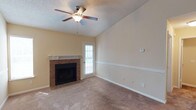 This single-floor space offers a comfortable and functional layout centered around a welcoming living area. The room is anchored by a classic fireplace framed in rich tile surround with a clean white mantel, providing a warm focal point for the space. Soft, neutral-toned carpet covers the floor, adding warmth and comfort underfoot, while the walls are finished in a light beige tone that creates a bright and airy atmosphere. A textured ceiling adds dimension overhead, complemented by a ceiling fan that ensures year-round comfort. Natural light filters into the room through a door fitted with blinds, which provides direct access to an outdoor area, offering both ventilation and a connection to the exterior. Decorative chair rail molding lines the walls, lending a subtle architectural detail that adds character to the room. The overall ambiance is clean, neutral, and move-in ready, with practical features thoughtfully integrated throughout. The space presents a straightforward and versatile layout suitable for a variety of furnishing arrangements and living styles.
