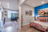 This single-floor unit is thoughtfully designed, blending modern finishes with bold, eclectic style. The kitchen is a standout feature, showcasing sleek two-tone cabinetry — high-gloss white upper cabinets paired with wood-grain lower cabinets — set against a gray tile backsplash. Stainless steel appliances, including a refrigerator, range, microwave, and dishwasher, complement the clean white countertops. A large window allows natural light to fill the space, offering views to the outside.

The open living area flows naturally from the kitchen and is anchored by a bold teal accent wall. Track lighting illuminates the room, highlighting eclectic furnishings that include a comfortable sofa, an industrial-style shelving unit, and an array of framed artwork. The space has a creative, gallery-like atmosphere.

The bedroom continues the vibrant teal accent wall, with recessed lighting and light wood-tone flooring found throughout. A closet provides practical storage, and the room exudes a cozy, artistic character.

The bathroom features a clean, modern design with a combination tub and shower, wood-grain cabinetry, and light tile flooring. A vivid orange door trim adds a pop of color to the hallway leading to the bathroom, tying into the unit's playful and distinctive design aesthetic.