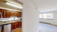 This single-floor unit offers a practical and efficient layout across three bedrooms and a kitchen. The kitchen is a standout feature, showcasing rich medium-toned wood cabinetry with ample upper and lower storage, granite countertops, a stainless steel single-basin sink, and a stainless steel electric range with an overhead exhaust hood. The kitchen floor features hardwood-style flooring, and fluorescent lighting keeps the space bright and functional. The living and bedroom areas are finished with wall-to-wall carpet in neutral tones, contributing to a warm, quiet atmosphere throughout. The spacious primary bedroom benefits from a wide window that brings in natural light and offers views of the surrounding building exterior, while a wall-mounted air conditioning unit and baseboard heating provide year-round climate control. A ceiling fan adds additional comfort to the living area. The remaining bedrooms are clean and simply finished, with carpeted floors, white walls, and dark wood interior doors that provide a classic contrast. The textured ceilings are consistent throughout the unit. The overall layout is straightforward and functional, with an open transition between the kitchen and living spaces, making efficient use of the available floor area.