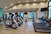 This fitness center offers a comprehensive and well-organized workout environment designed to accommodate a wide range of exercise routines. The space features vaulted ceilings with recessed lighting throughout, creating an open and energizing atmosphere. Large, black-framed windows line the walls, flooding the interior with natural light and providing views of the surrounding outdoor environment.

The main exercise area is generously proportioned and arranged into clearly defined workout zones. A row of Matrix elliptical trainers and upright cycling bikes occupies the central floor space, each unit equipped with its own individual display screen. Multiple treadmills are positioned along one side, offering ample cardio capacity. A dedicated strength and free weights area includes a dumbbell rack with a selection of weights and adjustable workout benches for versatile strength training.

Wall-mounted flat-screen televisions are positioned throughout the space for entertainment during workouts. A striking accent wall clad in teal subway tile adds a distinctive design element near the entry area, which also features a dual water fountain station and a paper towel dispenser for user convenience. The flooring throughout consists of durable, textured rubber-style material well suited for a high-activity environment. Glass interior partitions offer visibility between zones while maintaining an open, connected feel across the entire facility.