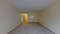 This single-floor space presents a straightforward and functional layout. The main living area is finished with wall-to-wall carpeting in a neutral gray tone, providing a clean and understated foundation. The walls are painted in a soft, warm off-white hue, contributing to a bright and open atmosphere. A notable architectural detail is the recessed rectangular niche built into the far wall, offering a dedicated spot for display or decorative purposes. The ceiling is smooth and finished in white, with a flush-mounted light fixture providing overhead illumination. Baseboards run along the perimeter of the room, adding a modest finishing detail. A hallway extends from the main living area, visible in the background and lit by an additional ceiling fixture, suggesting access to further rooms or utilities beyond the main space. The overall design is practical and neutral, making the space highly adaptable to a variety of furnishing styles and functional needs. The layout appears efficient, with clean sightlines throughout the main area.