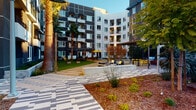 This vibrant outdoor courtyard serves as a central communal hub within a modern multi-story residential complex, showcasing a seamless blend of contemporary architecture and carefully curated landscaping. The space is framed by striking building facades that combine dark charcoal, crisp white, and bold terracotta-red cladding, creating a dynamic visual backdrop. Soaring palm trees anchor the courtyard, lending a warm, resort-like atmosphere, while string lights draped between posts add an inviting ambiance during evening hours.

The hardscaping is a standout feature, with geometric patterned pavers in alternating tones of light and dark laid in eye-catching chevron and grid formations, guiding visitors through the various zones of the courtyard. Lush, manicured turf areas provide open green space ideal for relaxation or casual recreation, complemented by native drought-tolerant plantings, ornamental grasses, and mulched garden beds that add texture and color throughout.

Dedicated social areas include shaded seating zones with bright yellow umbrellas, outdoor dining and lounge furniture, and a game table set on the turf. A covered pergola and bar-height seating further expand the entertainment options. Accessible walkways lead to a secured parking entrance, ensuring seamless connectivity. The overall ambiance is contemporary, lively, and welcoming.