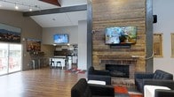 This single-floor space combines industrial and rustic design elements to create a warm, inviting communal environment. The open-concept layout allows for seamless flow between distinct functional zones, making it ideal for both relaxation and social gatherings.

The lounge area serves as a central focal point, anchored by a striking brick fireplace topped with a reclaimed wood mantel. The adjacent accent wall, clad in weathered wood planking, adds texture and warmth to the space, and a wall-mounted flat-screen television is positioned for comfortable viewing from the seating area below. Dark upholstered armchairs and a patterned area rug in bold red, grey, and black tones contribute to the cozy yet modern aesthetic.

Exposed structural beams and steel columns run through the space, reinforcing its industrial character, while a neutral grey and white color palette keeps the overall atmosphere clean and contemporary.

Toward the rear, a fully equipped kitchen features white cabinetry, a mosaic tile backsplash, and a central island with bar stool seating — a practical and stylish setup for food preparation and casual dining. A second wall-mounted television in this area ensures connectivity throughout the space. Hardwood-style flooring runs throughout, tying all areas together cohesively.