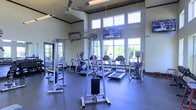 This fitness center is designed to support a comprehensive workout experience within a bright, open-concept layout. The space boasts soaring ceilings that create an airy, expansive atmosphere, complemented by a row of large windows along the far wall that flood the room with natural light and offer pleasant views of the surrounding greenery outside. A ceiling fan overhead aids in air circulation, contributing to a comfortable environment during workouts. The flooring consists of durable, dark rubberized material well-suited for high-traffic fitness use. The facility is outfitted with a robust selection of cardio equipment, including multiple treadmills and elliptical machines, providing ample options for cardiovascular training. Strength training is supported by a cable machine and additional weight equipment. Full-length mirrors line one wall, allowing users to monitor their form during exercise. Wall-mounted flat-screen televisions are positioned for easy viewing from the cardio equipment, adding an entertainment element to workout sessions. An exit door with glass paneling provides an additional source of natural light. Overall, the fitness center delivers a well-lit, fully functional, and inviting environment for a wide range of exercise routines.